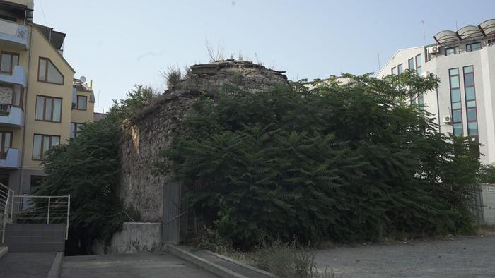The image on the left: Wide shot. The bathhouse Sredna Banya is concealed behind a thick wall of vegetation. Weeds and shrubs hairy the top of the dome, which peeks just above the vegetation that cordons it off. It is flanked on either side by modern apartment blocks. 