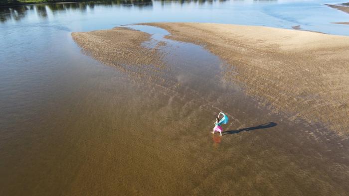 Woman performatively walking into a riverbed through shallow and sandy bank, holding behind a blue fabric with both of her hands, seen from an aerial perspective.