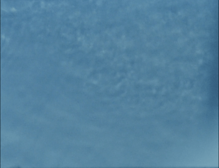A still image taken from 16mm film footage showing a swimming pool blue plastic tub being filled with water. The image is close up, and partly out of focus, showing a blue background and movement in the water. The still is taken from the film On Being and Bathing (2021) by Anna Ulrikke Andersen and Abi Palmer.  