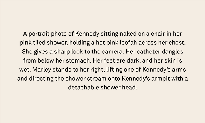 A portrait photo of Kennedy sitting naked on a chair in her pink tiled shower, holding a hot pink loofah across her chest. She gives a sharp look to the camera. Her catheter dangles from below her stomach. Her feet are dark, and her skin is wet. Marley stands to her right, lifting one of Kennedy’s arms and directing the shower stream onto Kennedy’s armpit with a detachable shower head.
