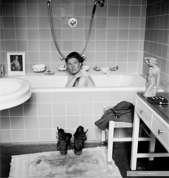A black and white photo partially shows a bathroom. In the background is a bathtub and Lee Miller is sitting right in the center. She is looking to the right. Her right hand scrubs her shoulder with a sponge. The shower is hanging on the wall, just above Lee Miller's head, forming a U. To the left, leaning against the wall, is a small portrait of Adolf Hitler from the torso up, wearing a military suit. On the floor are two black military boots facing the bathtub, and underneath, a small white rug is soiled with boot prints. To the right is a small white bench with an army uniform. Leaning against the bench is partially visible a white sideboard with a classical-style statuette of a nude woman, her right arm raised and resting on her head. The statuette faces in the direction of Lee Miller.