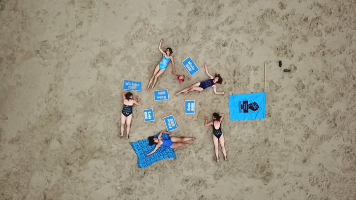 ive women in swimsuits, laying down on a sandy area, forming a visually striking composition with blue signs denoting river names and the flag representing the 'River Sisters' movement, seen from above.