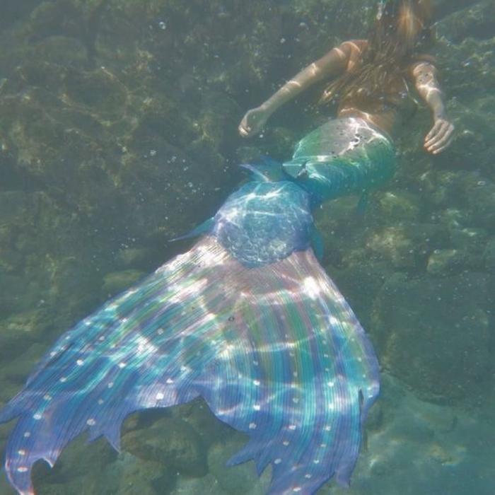 Person with long hair wearing a vibrant mermaid tail, seen from behind, floating in a rocky underwater setting.