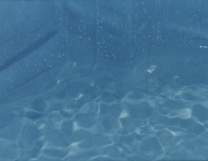 A still image taken from 16mm film footage showing a swimming pool blue plastic tub being filled with water. The image is close up, and partly out of focus, showing a blue background and movement in the water. The still is taken from the film On Being and Bathing (2021) by Anna Ulrikke Andersen and Abi Palmer.  