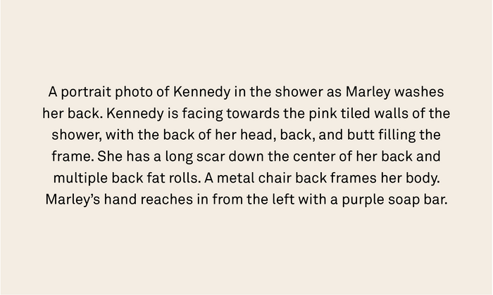 A portrait photo of Kennedy in the shower as Marley washes her back. Kennedy is facing towards the pink tiled walls of the shower, with the back of her head, back, and butt filling the frame. She has a long scar down the center of her back and multiple back fat rolls. A metal chair back frames her body. Marley’s hand reaches in from the left with a purple soap bar.