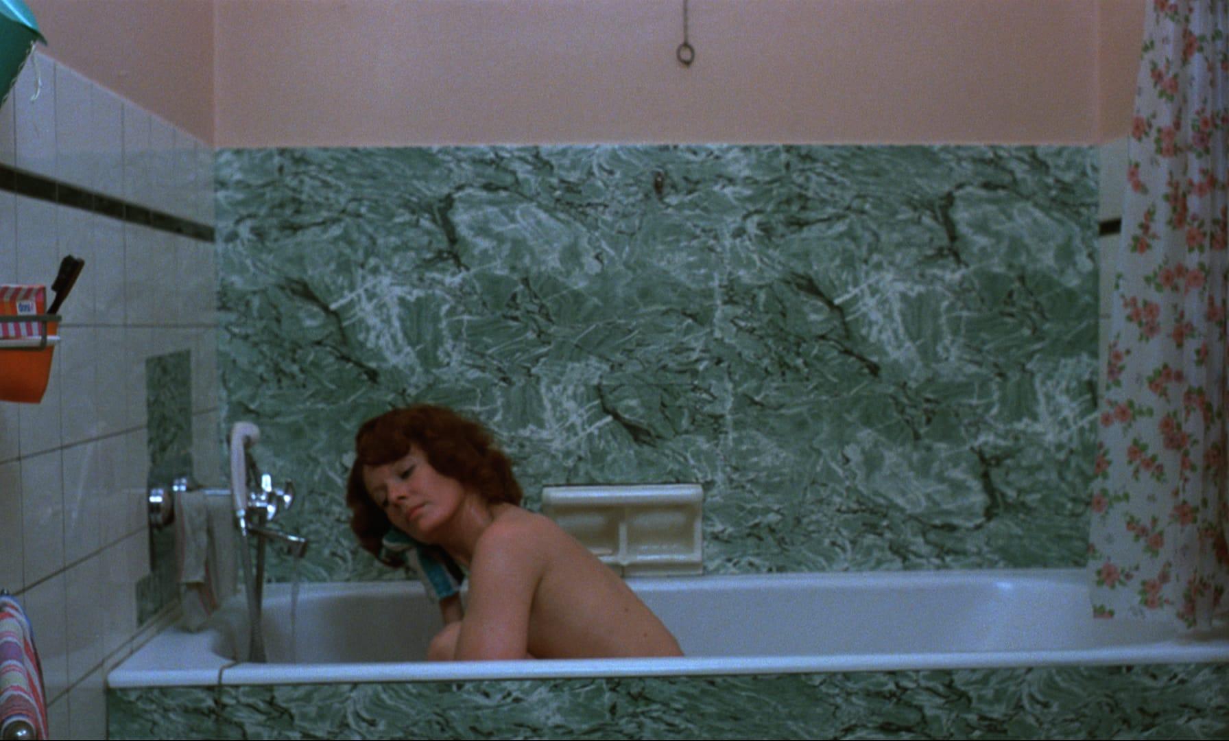A woman sitting in a spartan bathtub, rinsing her right ear.