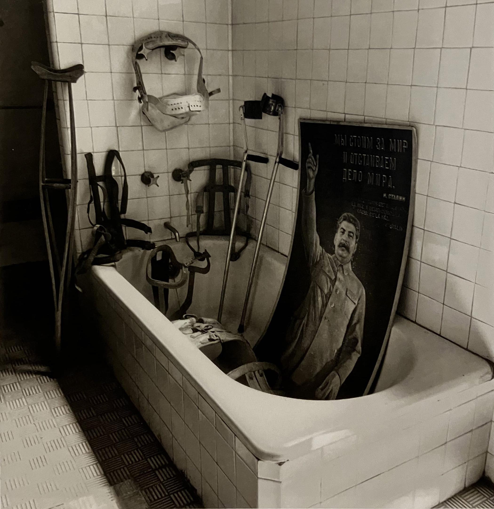 A black and white photograph of a white bathtub, a little bit dirty. The tiles on the wall are square and white. Clockwise, a big crutch is outside the tub, supported by the wall. Behind, on the tub, are some leather corsets. On the wall above, a used white plastic corset hung on two metal taps. Inside the bathtub, two crutches are leaning against the wall. Next to it a huge Soviet portrait of Joseph Stalin. In a military corporal expression, he raises his right arm, pointing upwards with his finger. There is a large plastic corset inside the bathtub, but we can't see the whole of it, only the top.